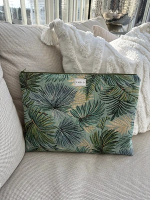 Grön laptopväska med palmbladsmönster - Snygg datorväska från EMELO med tropiskt palmbladsmönster i gröna och beige toner. Väskan har dragkedja upptill och är gjord i ett kraftigt tyg med vadderad insida. Perfekt för att bära din laptop på ett stilrent sätt. Passar laptop med maxmått: 33x24cm