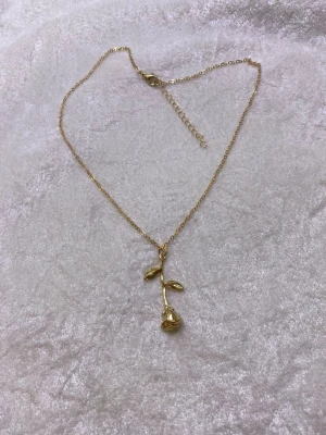 Guldigt halsband med rosberlock - Snyggt halsband i guldfärgad metall med en detaljrik rosberlock och två små blad. Kedjan är tunn och har justerbar längd. Perfekt accessoar för dig som gillar blommiga smycken och vill ha något unikt.