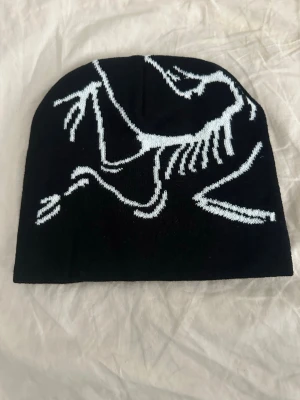 Mössa - Säljer en svart mössa med ett coolt vitt dinosaurieskelett-motiv. Mössan är stickad och har en enkel, klassisk form som passar perfekt till streetwear eller för att ge din outfit en unik touch.