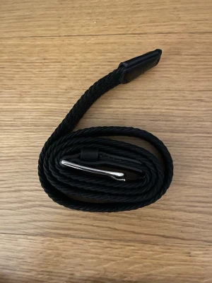 Flätat bälte - Tjenaritz! Ett snyggt svart flätat bälte med en stilren silverfärgad metallspänne. Bältet har en smal design och är gjort i syntetmaterial med detaljer i svart. Perfekt för att ge din outfit en clean och modern touch.