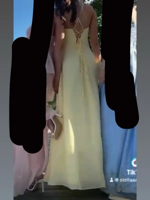 Balklänning -  Justerbar XS-S Elegant ljusgul långklänning med v-ringning, smala axelband och öppen rygg med snörningsdetalj. Klänningen har en slits framtill och är tillverkad i ett lätt, flytande material som ger en fin siluett. 