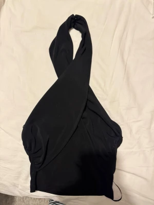 Svart halterneck topp - Snygg svart halterneck topp med omlott-design och breda axelband som korsas i nacken. Toppen är ärmlös och har en tight passform som framhäver figuren. Perfekt att styla med jeans eller kjol för en trendig look.