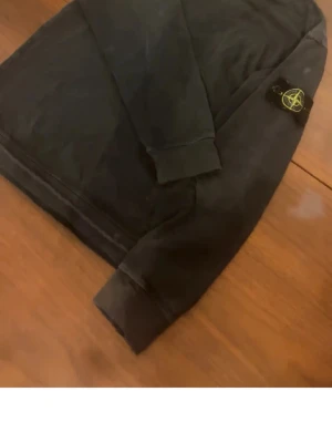 Svart sweatshirt från Stone Island - Säljer en svart sweatshirt från Stone Island med den klassiska loggan på ärmen. Tröjan har rund hals, ribbade muddar och är tillverkad i mjukt bomullsmaterial. Perfekt för dig som gillar streetwear och vill ha en stilren look.