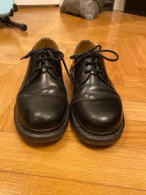 Svarta dr martens - Klassiska svarta loafers i blankt läder med rund tå och kraftig sula. Skorna har snörning framtill och en stilren design som passar till många olika outfits. Insidan är ljusbrun och sulan är robust för extra komfort.
