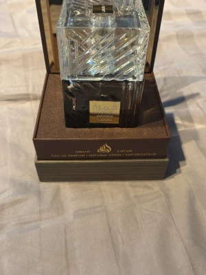 Lattafa Qahwa Eau de Parfum mot thurati - Lattafa Qahwa Eau de Parfum är en lyxig parfym i en elegant, fyrkantig glasflaska med detaljer i svart och guld. Flaskan har ett stilrent mönster och kommer i en exklusiv brun box. Perfekt för dig som vill ha en unik och so