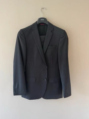 Hugo Boss Kostymset Svart - Säljer ett two-piece Kostymset från HUGO BOSS i Strl.46. Byxorna är custom sydda till en totallängd på 101cm. Färgen är svart med röda inner detaljer samt kritstrecks randiga ärmar på insidan. Den är som ny. Hör av dig vid frågor!