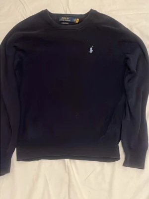Mörkblå tröja från Polo Ralph Lauren - Klassisk mörkblå tröja från Polo Ralph Lauren med rund halsringning och den ikoniska broderade loggan på bröstet. Tröjan har långa ärmar och är tillverkad i mjukt material som passar perfekt till många olika stilar.