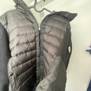 Svart pufferjacka från Moncler - Snygg svart pufferjacka från Moncler med stickade ärmar och vadderad front. Jackan har dragkedja framtill, hög krage och en liten Moncler-logga på ärmen. Perfekt för kalla dagar och riktigt trendig stil.