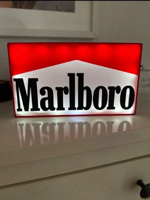 Marlboro ljuslåda skylt - Cool ljuslåda med Marlboro-logga i rött, vitt och svart. Skylten är rektangulär och tillverkad i plast, perfekt som dekoration eller samlarpryl. Den har inbyggd belysning som lyser upp loggan snyggt när den är tänd.