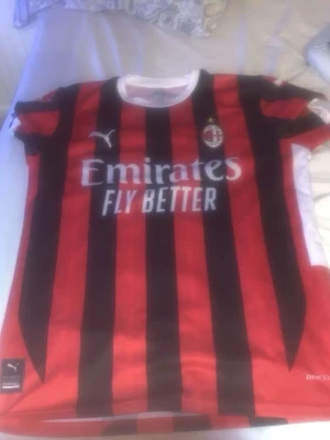 AC Milan matchtröja Ibrahimović 11 - Säljer en röd och svart AC Milan matchtröja från Puma med klassiska vertikala ränder. Framsidan har Emirates Fly Better-tryck och klubbmärke, baksidan har Ibrahimović och nummer 11. Perfekt för fotbollsfans som vill visa sitt stöd för laget.