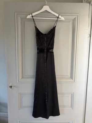 Svart långklänning från Zara - Elegant svart långklänning från Zara med smala axelband och knappar framtill. Klänningen har en lätt glansig yta och spetsdetalj vid midjan som ger en extra touch. Perfekt för dig som gillar stilrena och trendiga plagg.