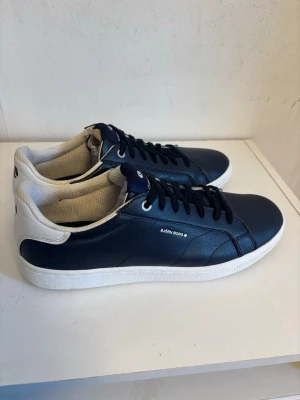 Marinblå sneakers från Björn Borg - Snygga marinblå sneakers från Björn Borg med vita detaljer och vit sula. Skorna har klassisk låg profil, svarta skosnören och ovandel i slätt konstläder. Perfekta för en clean och stilren look.