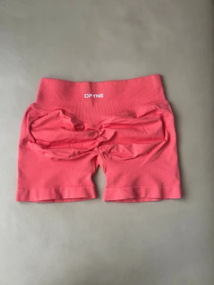 Korall träningstights shorts från DFYNE - Snygga rosa shorts från DFYNE med hög midja och tight passform. Tillverkade i ett stretchigt material som passar perfekt för träning. Shortsens baksida har en scrunch-detalj som framhäver formen. Bekväma och stilrena för gymmet eller löprundan.