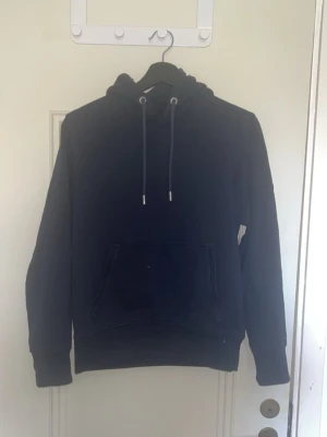 navy basic hoodie med ficka - En stilren svart hoodie med huva och dragsnören. Klassisk känguruficka framtill och ribbade muddar vid ärmslut och nederkant. Perfekt för en avslappnad look. Materialet känns mjukt och bekvämt, troligtvis bomullsblandning.
