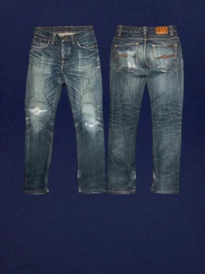 Nudie Jeans - Säljer ett par blå jeans från Nudie Jeans med slitningar och hål på framsidan samt slitna detaljer på bakfickorna. Klassisk femficksmodell med raka ben och tydliga kontrastsömmar. Tillverkade i denim med en cool tvättad look.