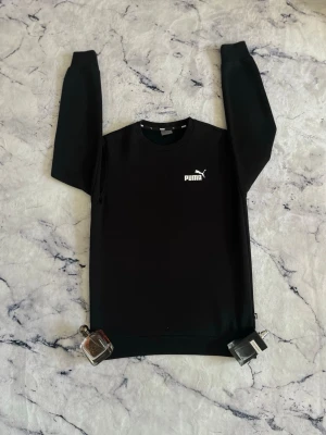 Svart sweatshirt från Puma - Svart sweatshirt från Puma med rund halsringning och vit Puma-logga på bröstet. Tröjan har en klassisk passform med långa ärmar och ribbade muddar vid ärmslut och nederkant. Perfekt för en sportig och avslappnad stil.