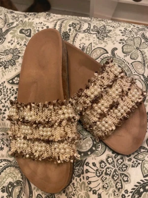 Beige sandaler med pärldetaljer - Säljer ett par beige sandaler med breda band dekorerade med pärlor och vävt tyg i bruna och krämiga toner. Sandalerna har en platt sula och öppen tå, perfekta för en trendig och avslappnad look. Unika detaljer som ger extra stilpoäng.
