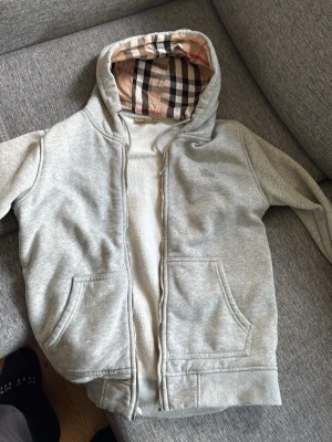Grå hoodie med Burberry-mönstrad huva - Snygg grå hoodie med dragkedja och klassiskt Burberry-rutigt foder i huvan. Två stora fickor framtill och ribbade muddar vid ärmslut och nederkant. Perfekt för en avslappnad och trendig stil.