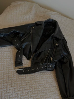 ZARA Svart skinnjacka med bälte och dragkedjor  - Snygg svart skinnjacka från Zara med klassisk bikerstil, silvriga dragkedjor och bälte i midjan. Jackan har bred krage och flera fickor med dragkedja för en cool look. Perfekt för dig som vill ha en edgy stil.