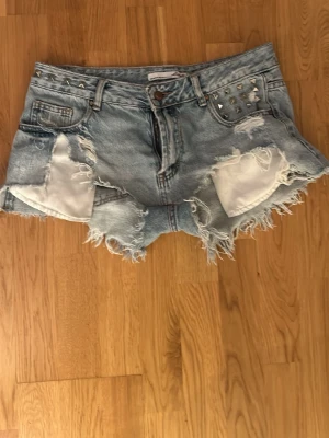 Rippade lågmidjadd jeansshorts med nitar - Snygga ljusblå jeansshorts med slitna detaljer och fransig kant. Shortsen har nitar på ena fickan fram och bak, samt klassisk femficksmodell. Perfekta för en cool och avslappnad stil. Köpta här på Plick, omsydda för att de ska sitta mer lågmidjat, passar S-M