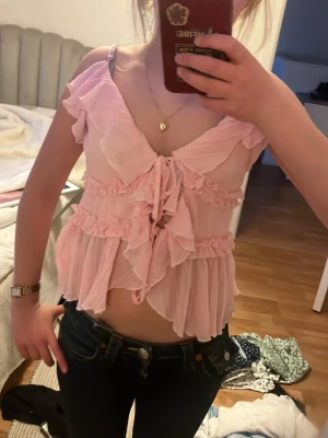Rosa volangig blus med knyt fram - Supersöt ljusrosa blus med volanger och knyt fram. Blusen är ärmlös med tunna axelband och har en luftig, omlottliknande design.