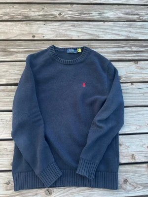 Mörkblå stickad tröja Polo Ralph Lauren - Mörkblå stickad tröja från Polo Ralph Lauren med klassisk rund halsringning och röd broderad logga på bröstet. Tröjan har ribbade muddar vid ärmslut och nederkant, perfekt för en stilren och enkel look.