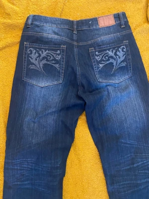 Mörkblå jeans med broderade bakfickor - Snygga mörkblå jeans med dekorativa broderier på bakfickorna. Jeansen har klassisk femficksmodell och subtila slitningar framtill för en cool look. Perfekta för dig som gillar detaljer och vill sticka ut lite extra.