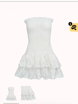 Vit axelbandslös volangklänning - Supersöt vit kortklänning med smockad överdel och volangig kjol i broderad bomull. Axelbandslös modell som ger en fräsch sommarkänsla, perfekt för varma dagar. Klänningen har fina spetsdetaljer och dubbla volanger nertill.