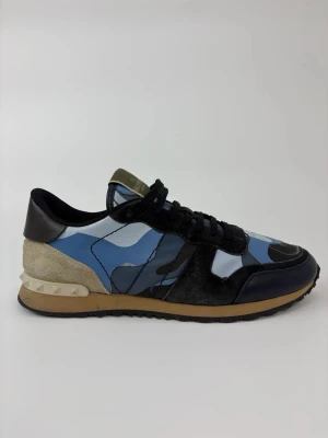 Valentino blå camo sneakers - Snygga sneakers från Valentino med blått camo-mönster, svarta detaljer och beige mocka vid hälen. Skorna har svarta skosnören, gummisula i beige och svart samt nitar på häldelen. Materialmix av läder, mocka och textil ger en lyxig känsla.