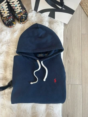 Mörkblå hoodie från Polo Ralph Lauren - Snygg mörkblå hoodie från Polo Ralph Lauren med vit dragsko och klassisk röd logga broderad på bröstet. Hoodien har en stor huva och är tillverkad i mjukt bomullsmaterial, perfekt för en avslappnad och stilren look.