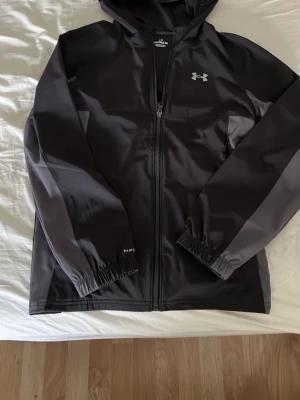 Svart träningsjacka från Under Armour - Svart träningsjacka med huva från Under Armour. Jackan har hel dragkedja, elastiska muddar och diskret logga på bröstet. Tillverkad i ett lätt och funktionellt material som passar perfekt för sport och träning.