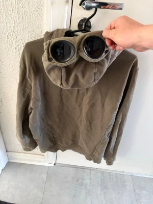 Olivgrön hoodie med goggles CP Company - Säljer en olivgrön hoodie från CP Company med dragkedja och två fickor framtill. Huvan har inbyggda goggles som ger en unik och cool look. Perfekt för dig som vill sticka ut med streetwear-stil. Materialet känns mjukt och bekvämt.