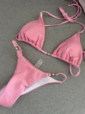 Rosa bikini med struktur och pärldetaljer - Snygg rosa bikini från SHEIN med strukturerat tyg och fina guldfärgade pärldetaljer på både över- och underdel. Klassisk trekantsmodell med knyt i nacken och ryggen. Perfekt för strandhäng och poolparty!