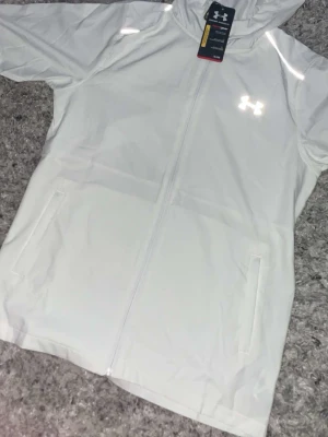 Vit vindjacka från Under Armour - Snygg vit vindjacka från Under Armour med huva och hel dragkedja. Jackan har två fickor framtill och reflekterande logga på bröstet samt stort reflekterande UNDER ARMOUR-tryck på ryggen. Perfekt för blåsiga dagar och sportiga outfits.