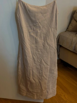 Beige klänning från H&M - Stilren beige långklänning från H&M med smala axelband och rak passform. Klänningen är ärmlös och har en enkel, minimalistisk design som passar perfekt till varmare dagar. Tillverkad i ett mjukt material som känns skönt mot huden.