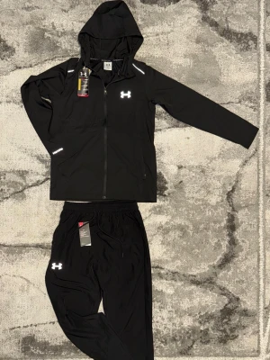 Svart sett från Under Armour - Snyggt svart sett från Under Armour med jacka och byxor. Jackan har huva, hel dragkedja och reflekterande detaljer samt logga på bröstet. Byxorna har resår i midjan, dragsko och logga på benet. Perfekt för löpning eller gym.