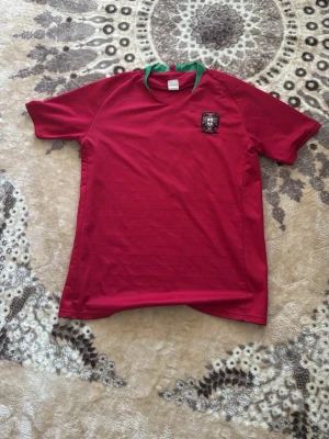 Portugal fotbollströja Ronaldo 7 - Röd Portugal fotbollströja med gröna detaljer i kragen och landslagets emblem på bröstet. På ryggen står det 'Ronaldo' och nummer 7 i guld. Perfekt för dig som vill stötta Portugal eller är ett fan av Ronaldo.