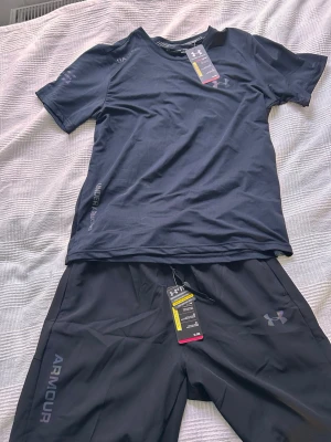 Svart träningsset från Under Armour - Snyggt svart träningsset från Under Armour med t-shirt och shorts. Båda plaggen har diskreta loggor och är gjorda i ett lätt, snabbtorkande material som andas. Perfekt för gymmet eller löprundan. Sportig och modern design med bekväm passform. Står storlek L men sitter som M
