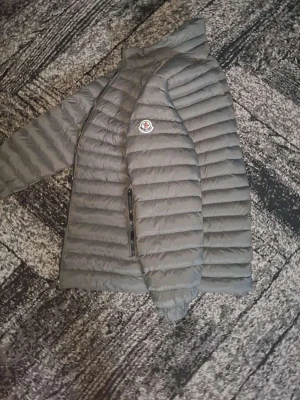 Grå dunjacka från Moncler - Snygg grå dunjacka från Moncler med quiltad design och raka kanaler. Jackan har två dragkedjeförsedda fickor framtill och en hög krage. Moncler-logga på vänster ärm. Perfekt för kalla vinterdagar.