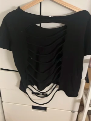 Svart topp med öppen rygg och snörning - Cool svart topp med kort ärm och öppen rygg med snörningsdetaljer. Toppen är croppad och har en avslappnad passform. Perfekt för dig som vill sticka ut med en edgy stil.