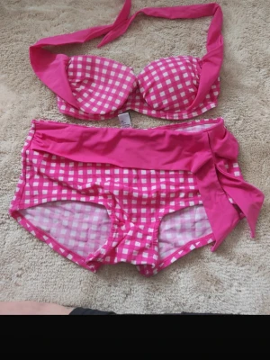 Rutig rosa bikini - Supersöt bikini från Swimwear by ellos i rosa och vitt rutigt mönster. Överdelen har halterneck och vadderade kupor, och nedre delen har en bred knyt i sidan för extra retrovibe. Perfekt för strandhäng eller poolparty! Passar s