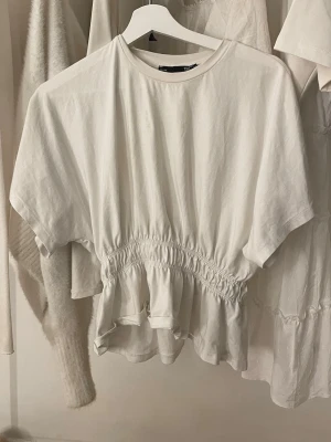 Vit topp med smock och volang - Vit topp från H&M med korta ärmar och smockad midja som ger en snygg volangdetalj nedtill. T-shirten har en rund halsringning och är gjord i mjuk bomull, perfekt för en avslappnad och trendig look. Xs/S