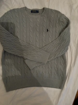 Ljusgrå kabelstickad tröja Polo Ralph Lauren - Snygg ljusgrå kabelstickad tröja från Polo Ralph Lauren med rund halsringning och broderad logga på bröstet. Tröjan har ribbade muddar vid ärmslut och nederkant, perfekt för en stilren och klassisk look.