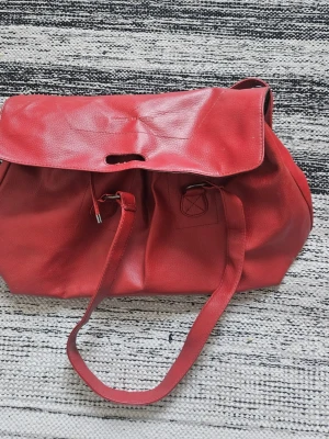 Röd ryggsäck i skinnimitation - Snygg röd ryggsäck i skinnimitation med dragsko och lock. Rymlig insida med mörkt foder och innerfack. Justerbara axelband och enkel design som passar till många stilar. Perfekt för dig som vill ha en färgklick till din outfit.