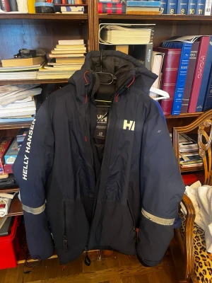 Marinblå Helly Hansen vindjacka - Snygg marinblå vindjacka från Helly Hansen med huva, dragkedja och reflexdetaljer på ärmarna. Stor logga på bröstet och ärmen. Jackan har flera fickor med dragkedja och justerbar nederkant. Perfekt för blåsiga dagar och outdoor-äventyr. Drag kedjan har hoppat ur  spåret men går att fixa.