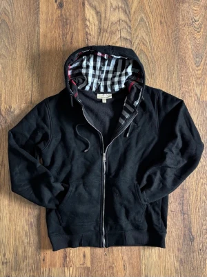 Svart hoodie från Burberry M - Storlek L men passar som M! Snygg svart hoodie från Burberry med dragkedja och klassiskt rutigt foder i huvan. Hoodien har fickor framtill och justerbar dragsko i huvan. Perfekt för dig som gillar stilrena plagg med exklusiv känsla.