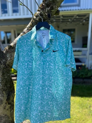 Golf pike från Nike - Fräsch mintgrön t-shirt från Nike med vitmönstrat tryck och klassisk svart swoosh-logga på bröstet. T-shirten har krage och två knappar framtill, perfekt för en sportig och avslappnad stil.