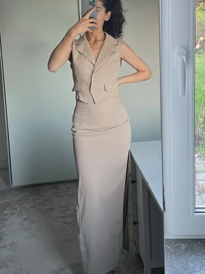 Kjol och tröja set  - Stilren beige byxdress med ärmlös västtopp och bred krage. Byxdressen har knappar framtill, dekorativa fickor och en lång, figurnära nederdel med hög slits på sidan. Perfekt för dig som gillar minimalistisk och trendig stil.