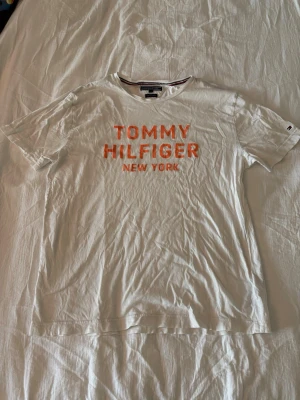 Vit T-shirt Tommy Hilfiger XL - Vit T-shirt från Tommy Hilfiger med orange text 'TOMMY HILFIGER NEW YORK' på bröstet. Tillverkad i ekologisk bomull och har klassisk passform med korta ärmar. Perfekt för en avslappnad och stilren look.