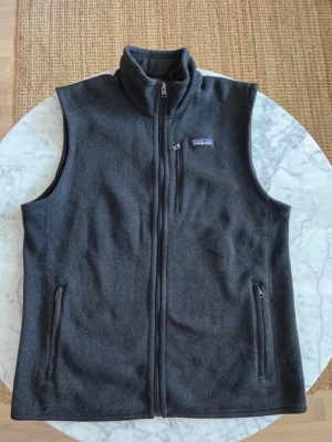 Mörkblå fleeceväst från Patagonia - Mörkblå väst i mjuk fleece från Patagonia med hel dragkedja framtill, två sidofickor med dragkedja och en bröstficka. Hög krage och enkel, sportig design som passar perfekt till lager-på-lager-stil.